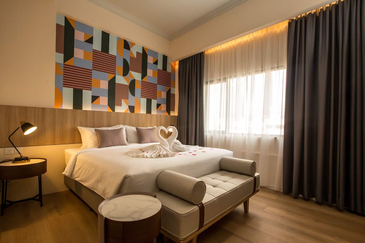 A modern hotel room with a large bed, a bedside table with a lamp, a decorative wall art piece, and a view of the outdoors through sheer curtains.