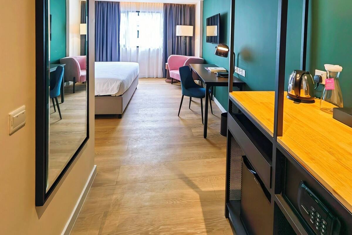 A hotel room with a bed, a desk with a computer, a chair, a mirror, and a coffee maker.