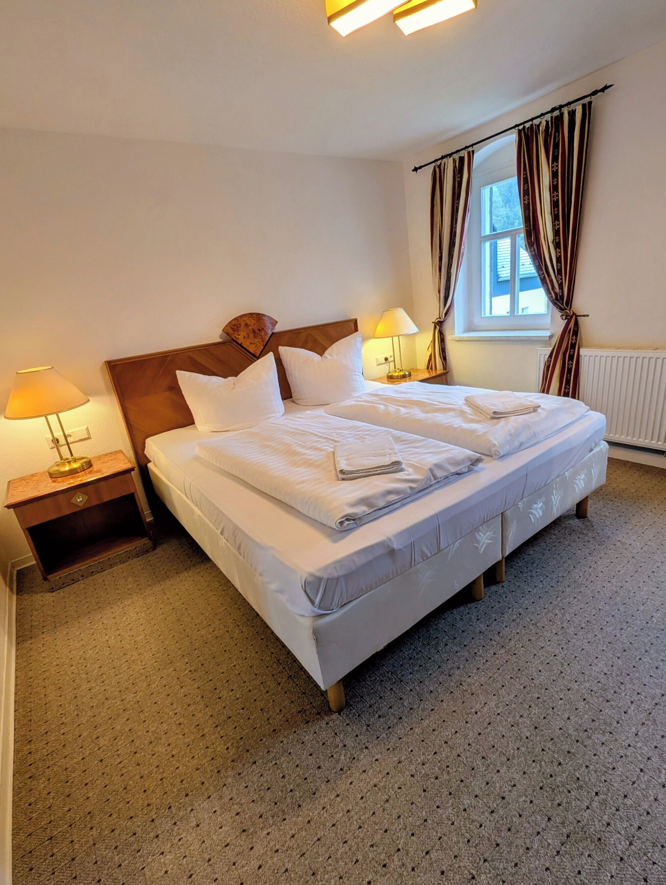Une chambre d’hôtel avec un lit double, deux tables de chevet avec des lampes, une fenêtre avec des rideaux et un radiateur.
