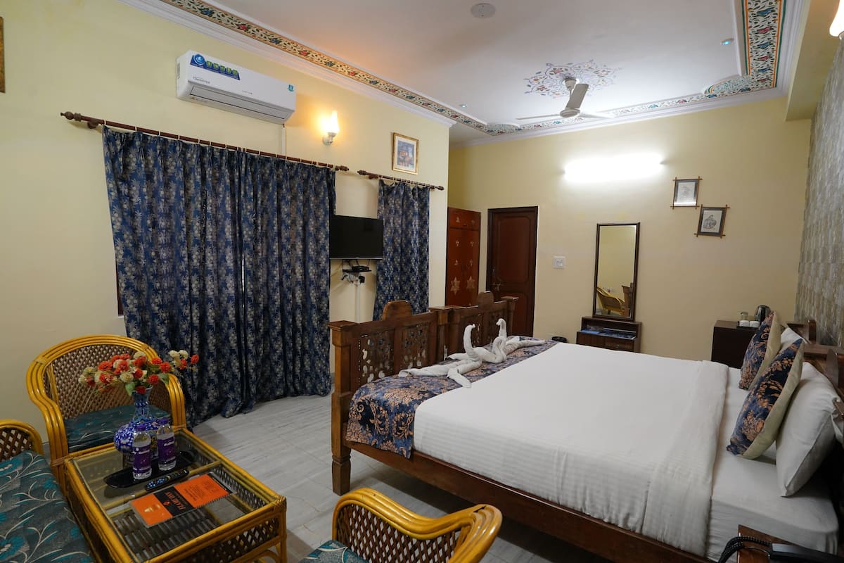 Une chambre d’hôtel comprenant un lit, une télévision, un ventilateur de plafond, une fenêtre avec des rideaux et une table sur laquelle se trouvent un vase et des bouteilles.