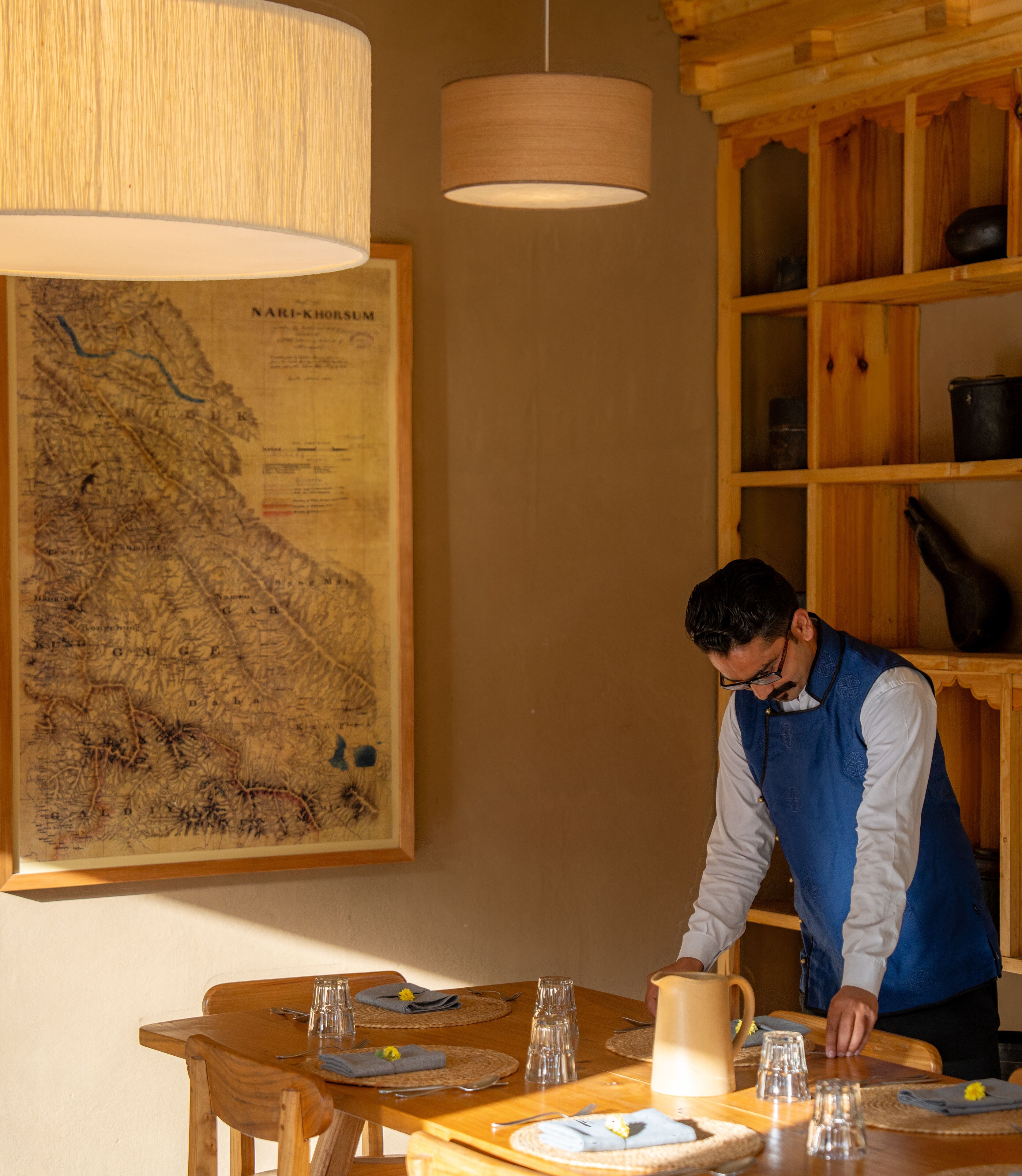 A person setting a table with a map and wooden furniture in a room with a wooden cabinet and hanging lamps.