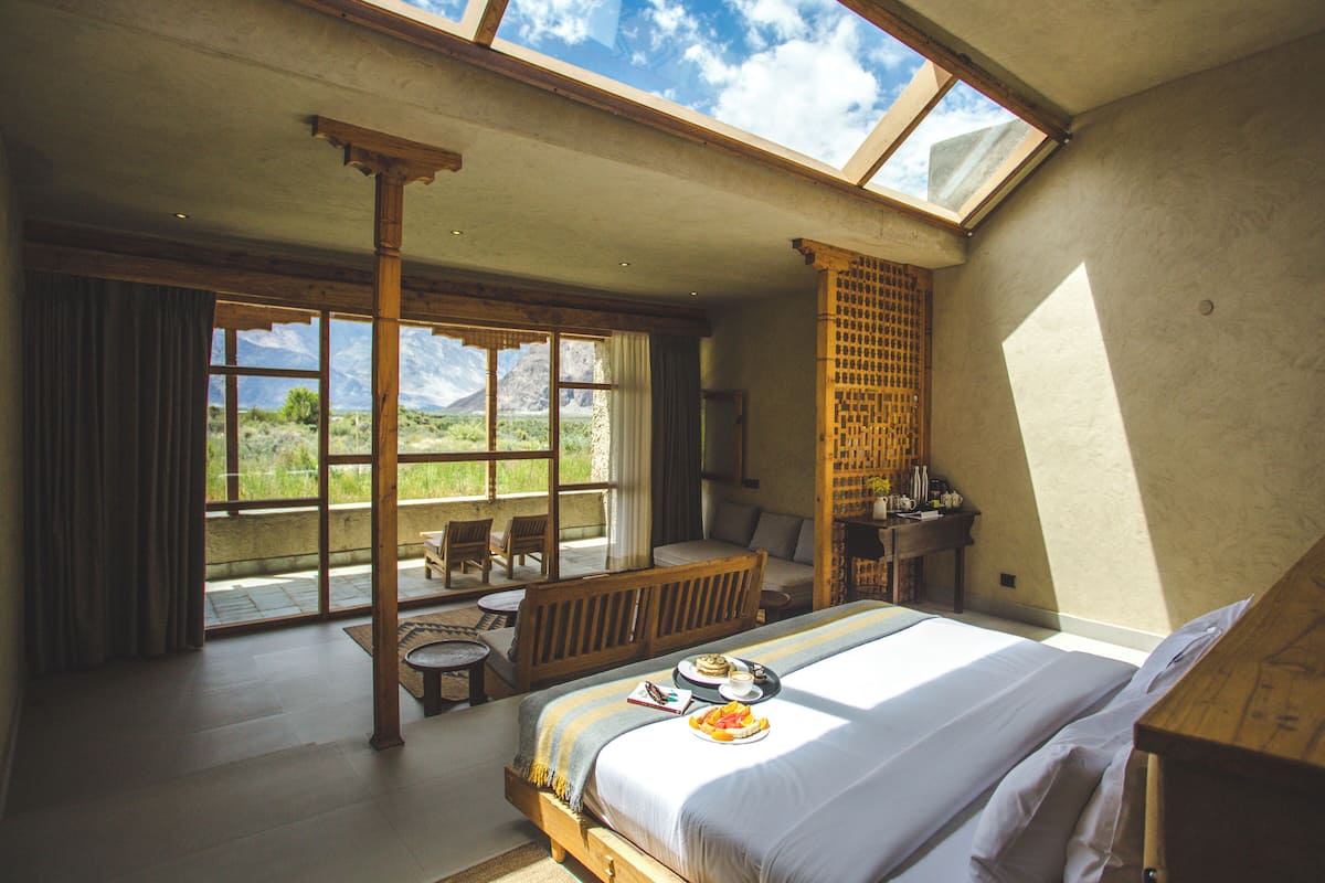 Une chambre avec un grand lit, un banc en bois, une petite table et une vue sur les montagnes par de grandes fenêtres.