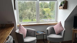Une pièce chaleureuse avec deux fauteuils gris, une petite table, un radiateur et une grande fenêtre donnant sur un paysage verdoyant.