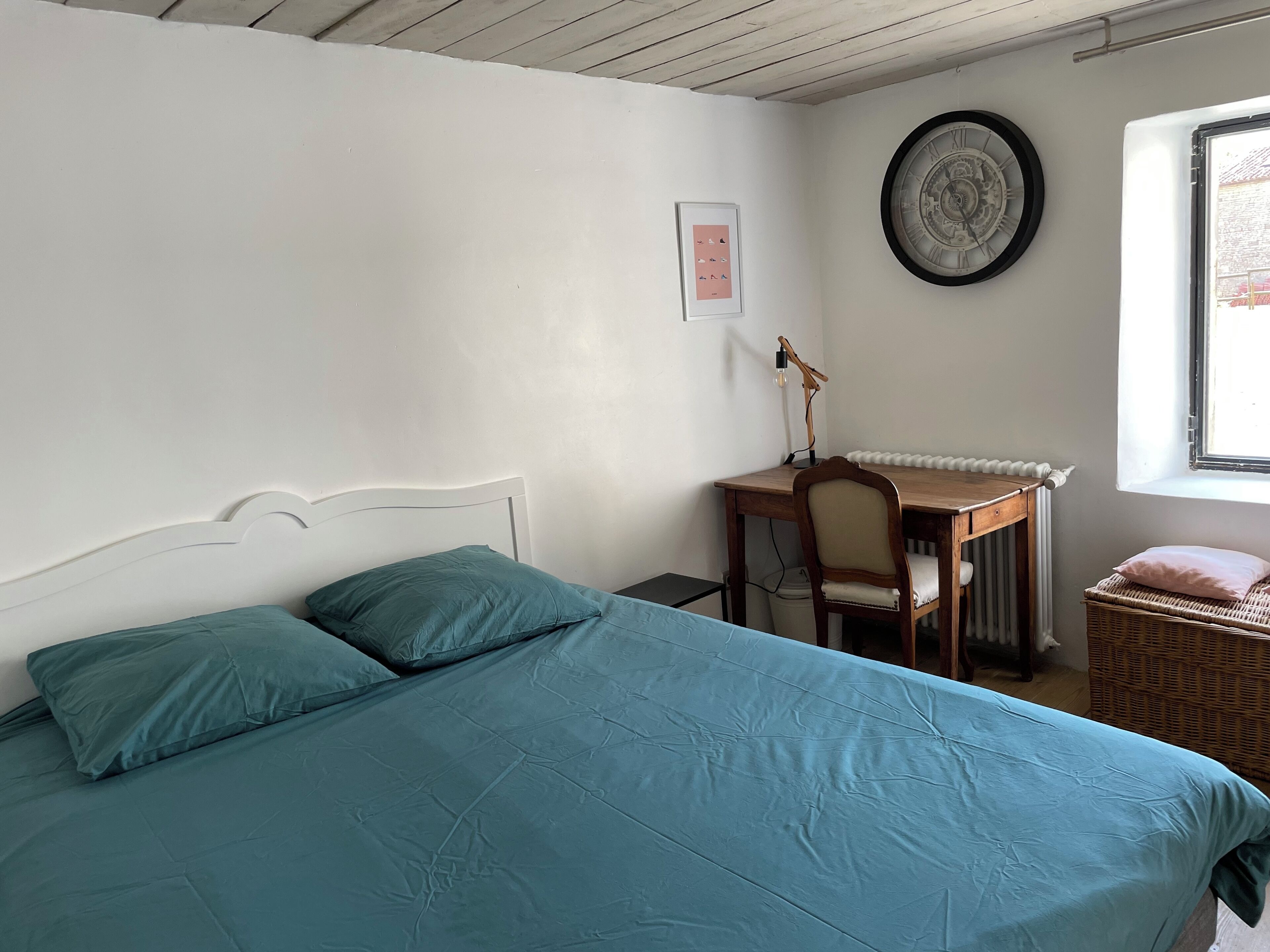 A bedroom with a bed, a desk, a chair, a wicker chair, a radiator, and a window.