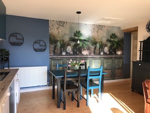 A dining area with a blue wall, a mural with tropical plants, a white radiator, and a wooden table with blue chairs.