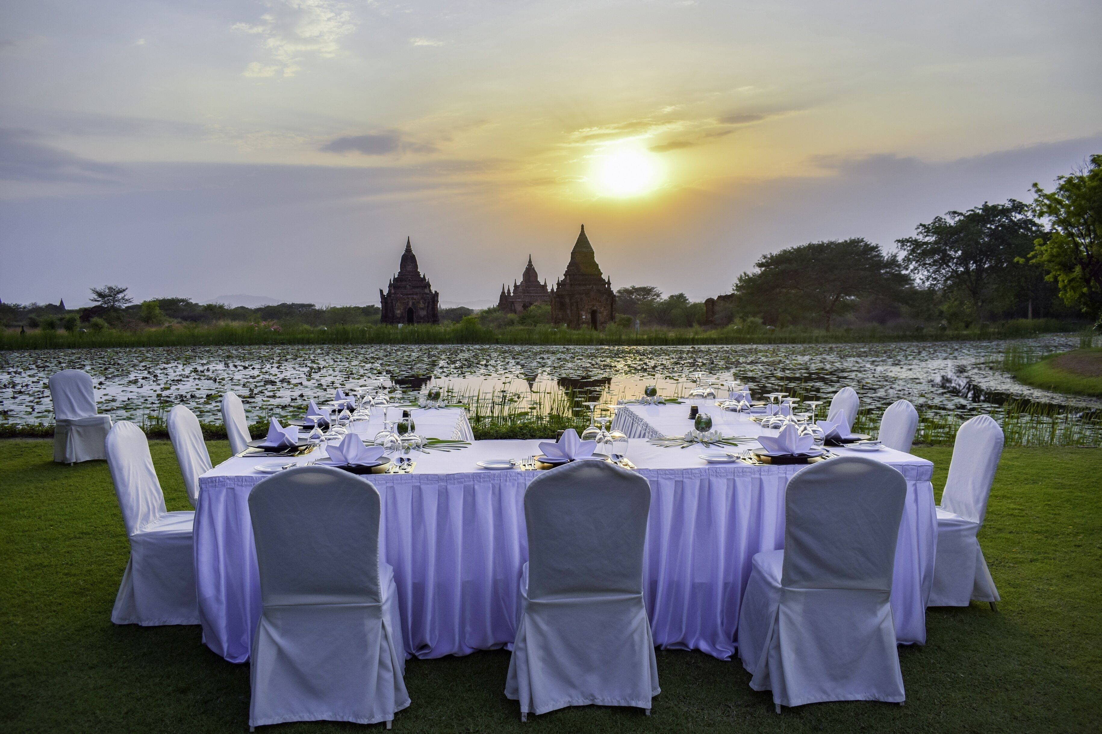 A table set for a meal with a view of temple ruins and a sunset over a body of water.