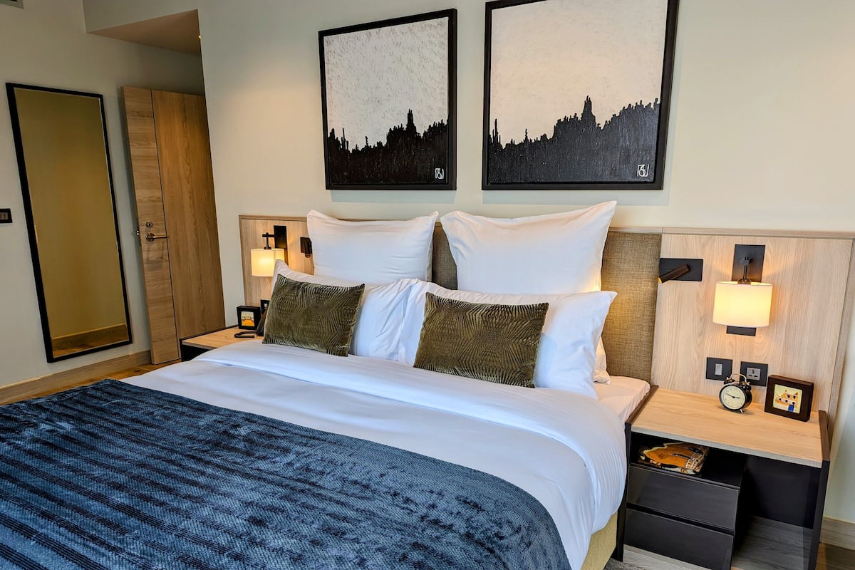 A neatly made bed with white linens and two green pillows, a bedside table with a clock and a picture frame, and two wall-mounted lamps.