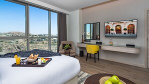 A hotel room with a large bed, a desk with a chair, a television, and a view of the desert landscape. - Citadines Abha (Abha)