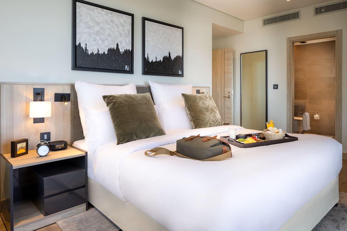 A hotel room with a large bed, a bedside table with a lamp, a nightstand, and a tray with breakfast items.
