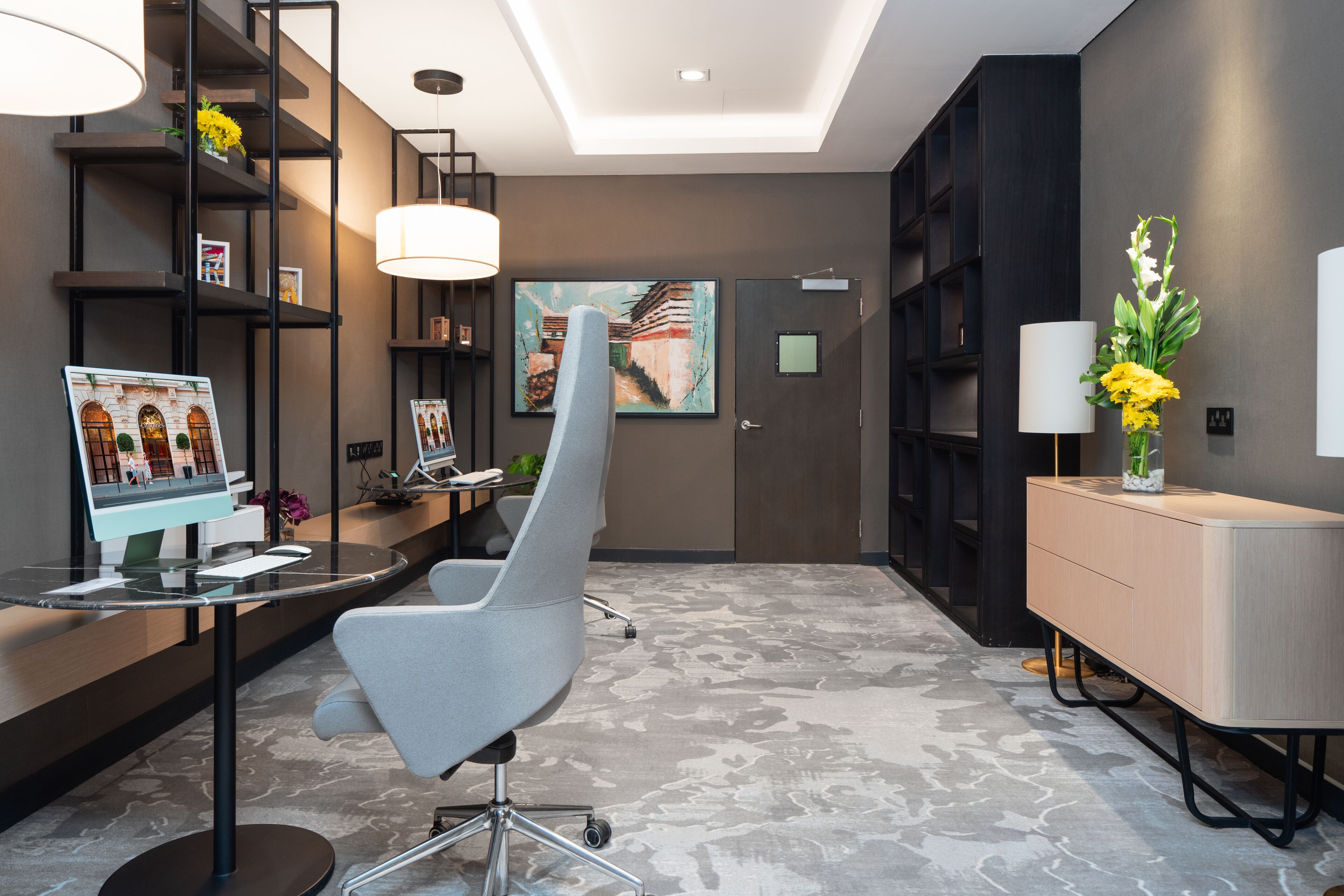 A modern reception area with a desk, a grey chair, a large painting, and a bookshelf.