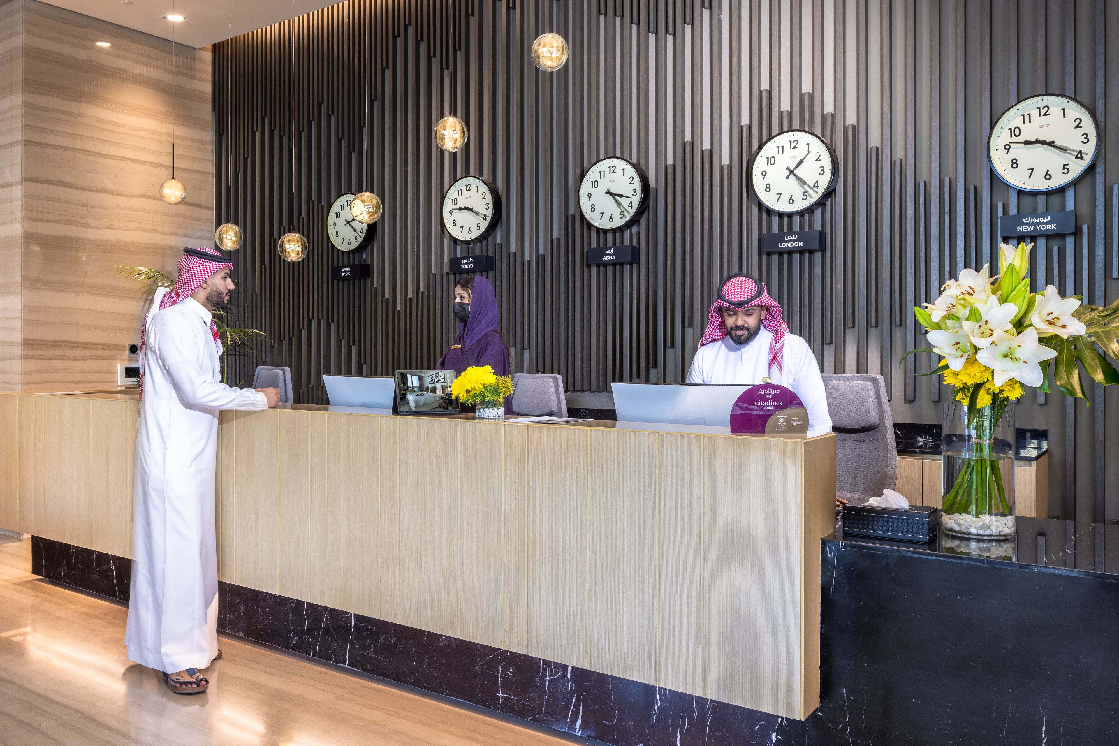 A hotel reception desk with multiple clocks displaying different time zones, a modern interior with vertical wooden panels, and a vase of flowers on the right.
