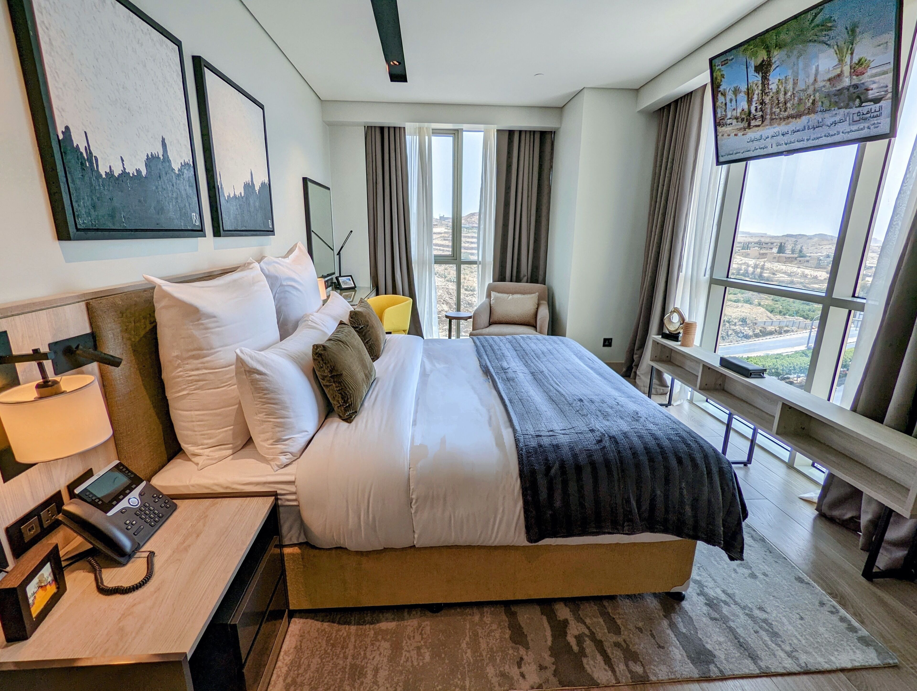 Una habitación de hotel moderna con una cama grande, teléfono junto a la cama y vistas a la ciudad a través de amplios ventanales.