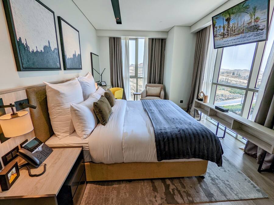 Una habitación de hotel moderna con una cama grande, teléfono junto a la cama y vistas a la ciudad a través de amplios ventanales.