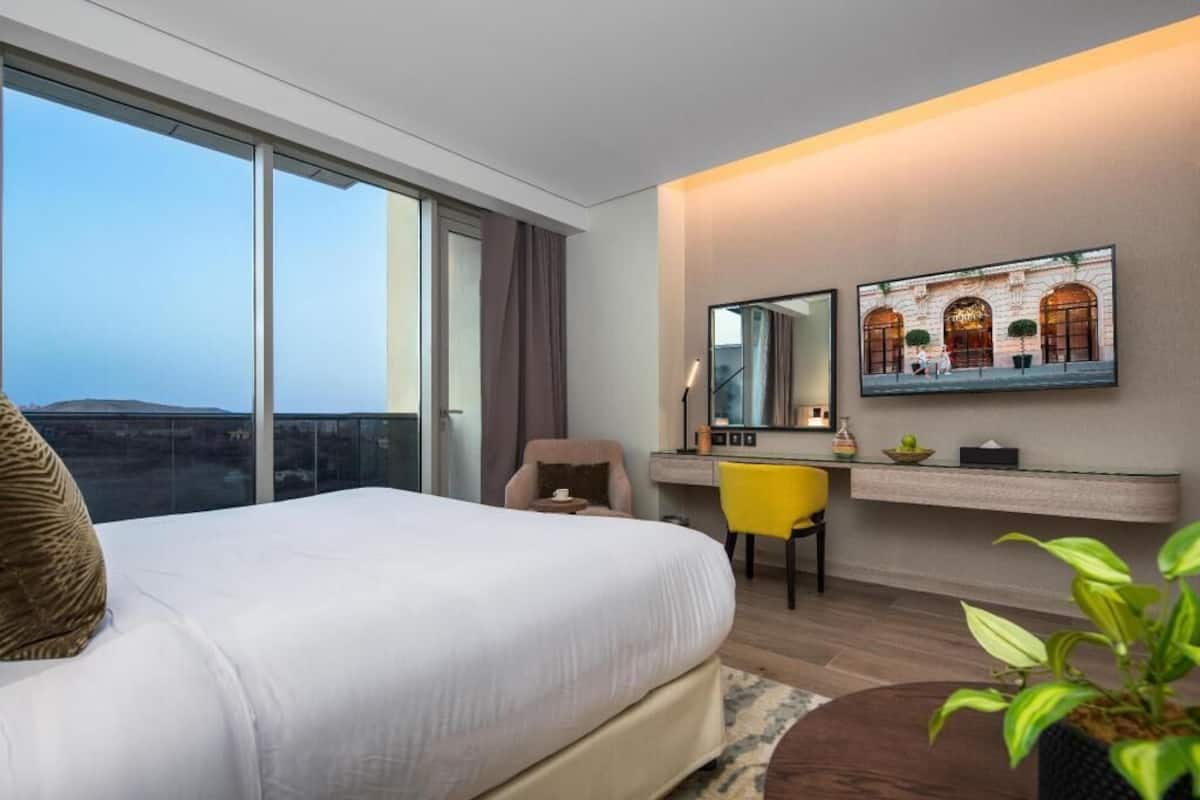 A hotel room with a large bed, a desk, a chair, a mirror, and a view of the cityscape.