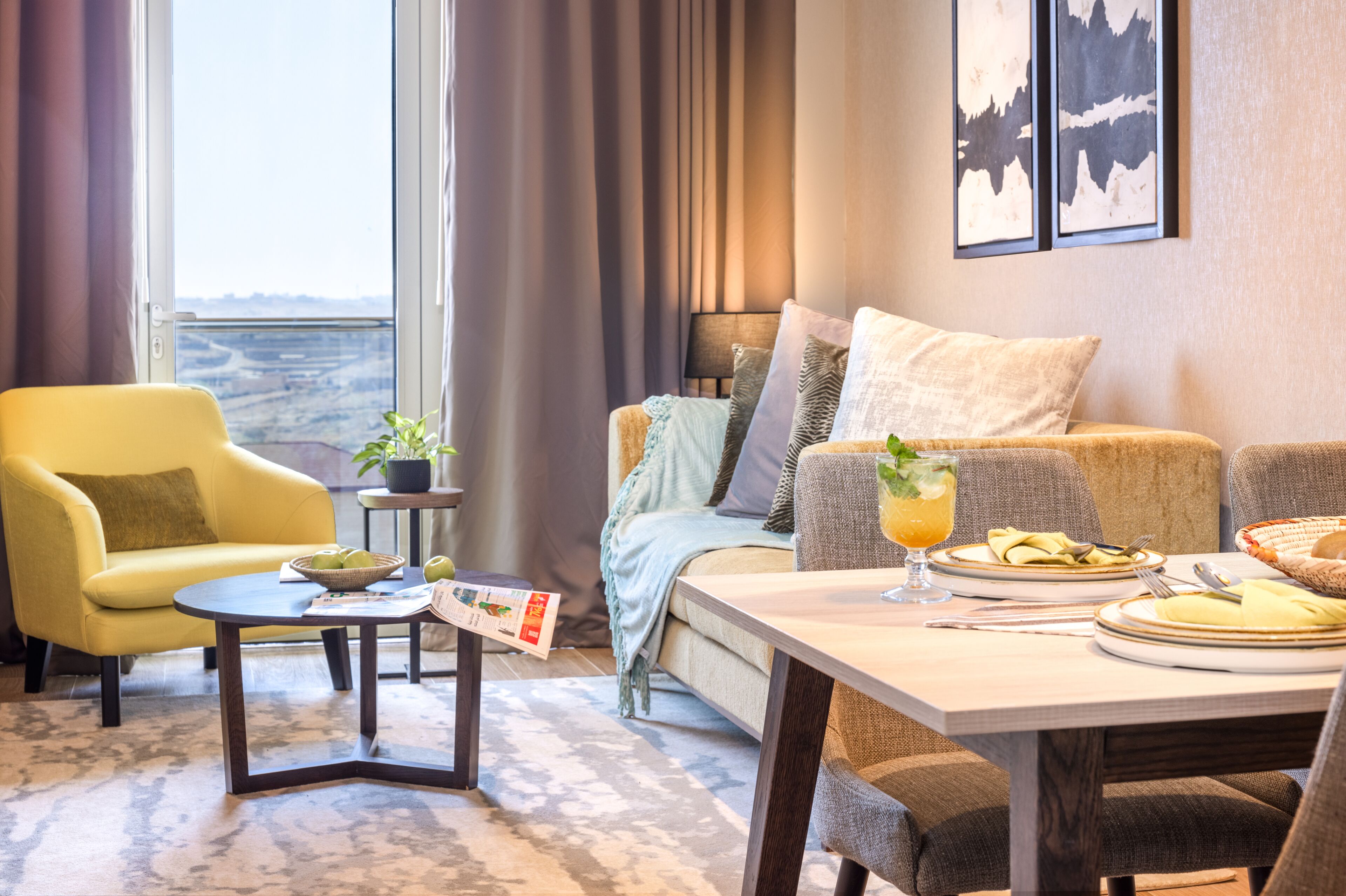 Ein gemütliches Wohnzimmer mit einem gelben Sessel, einer Couch, einem Esstisch für zwei Personen und einem Blick ins Freie durch große Fenster.