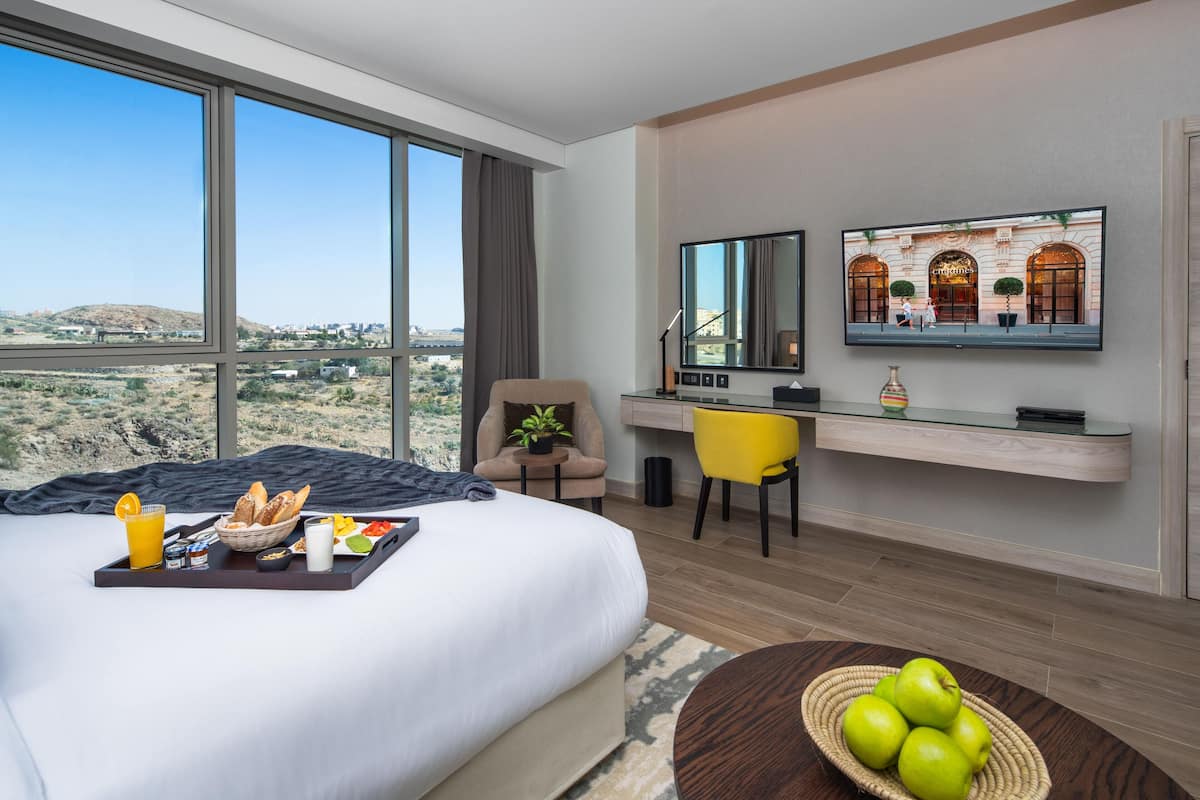A hotel room with a large bed, a desk with a chair, a television, and a view of the desert landscape.