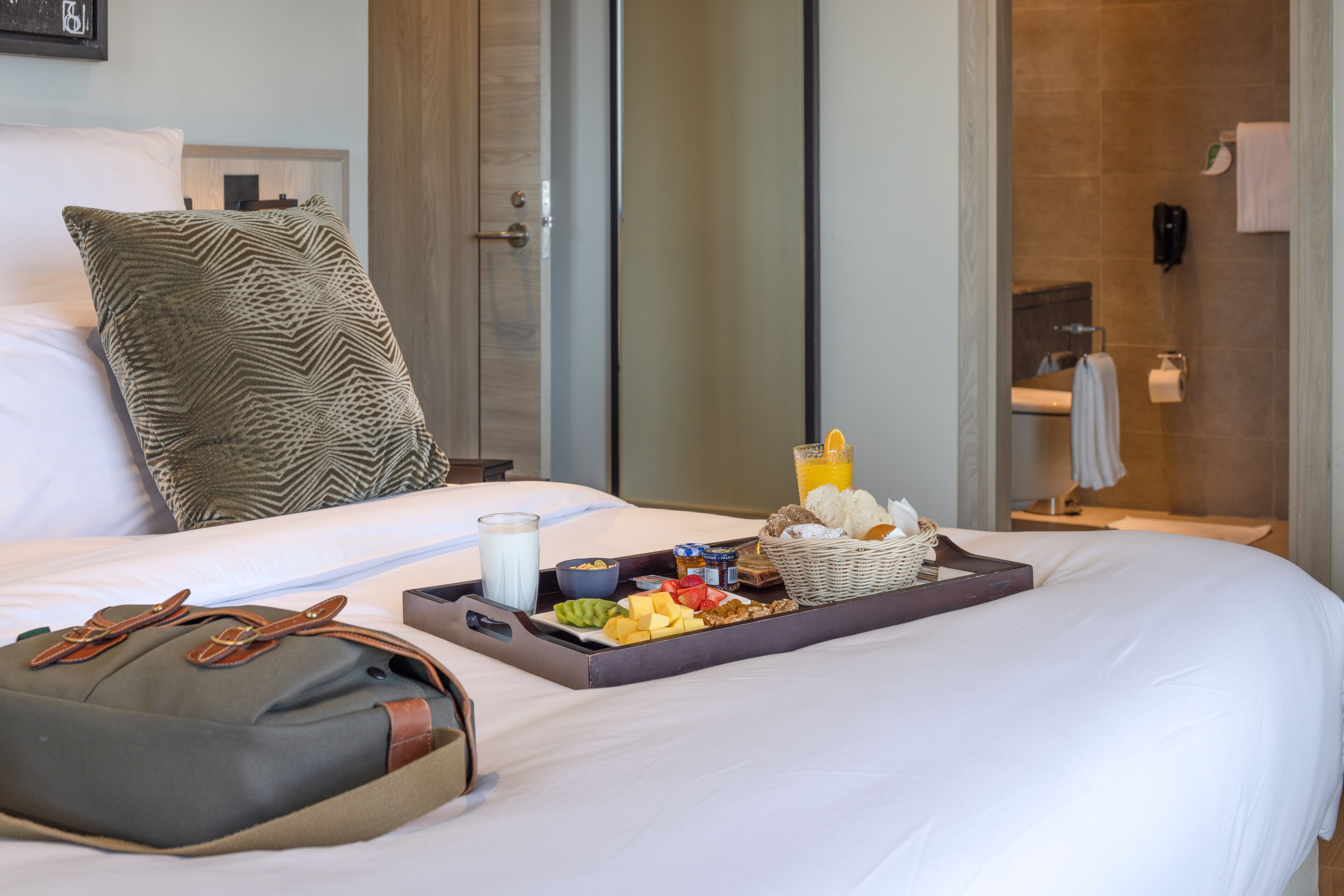A neatly made bed with a tray of breakfast items, a pillow, and a bag on the side.