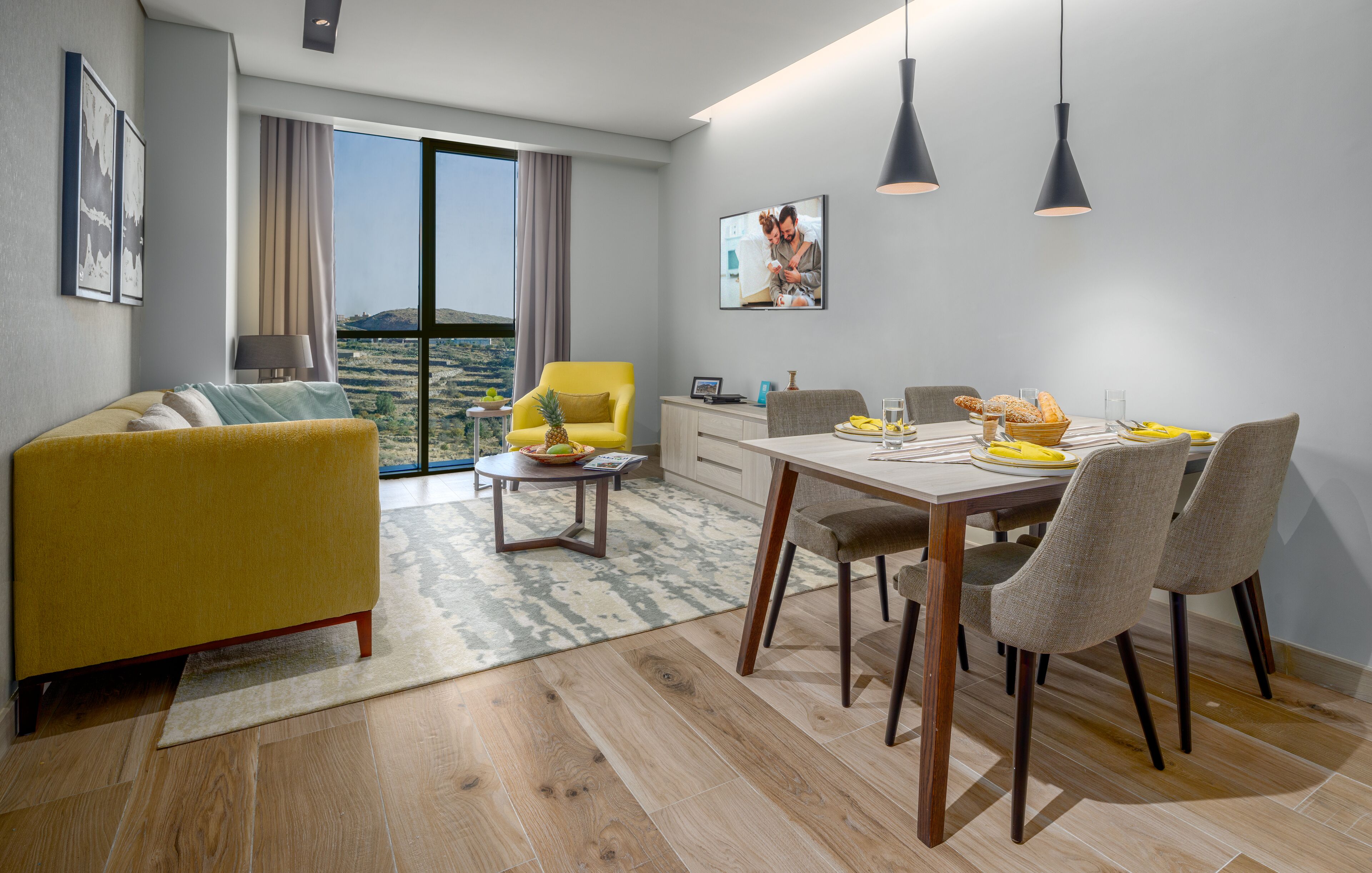 A modern living room with a yellow sofa, dining table, and chairs, a small coffee table, and a view of the outdoors.