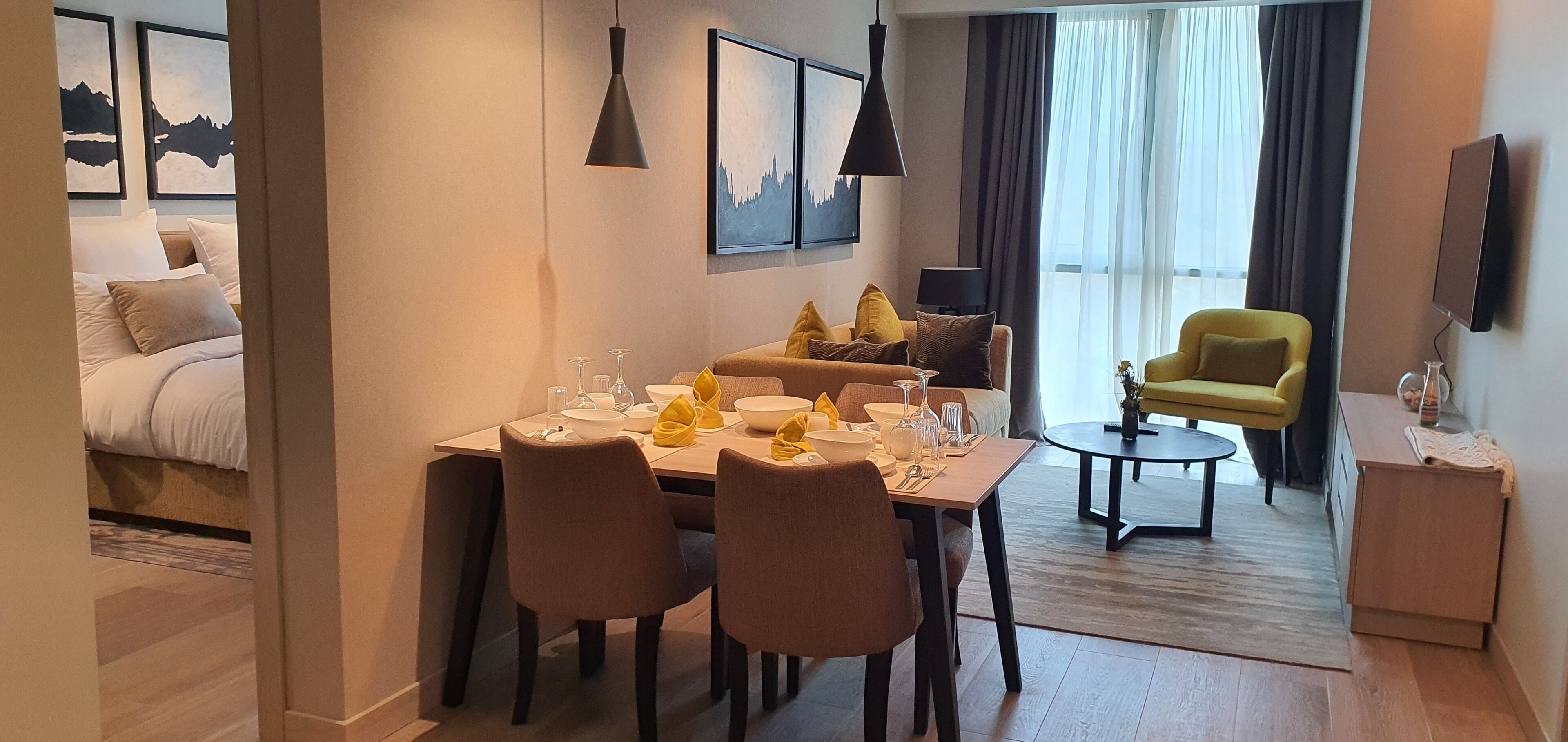 A modern hotel room with a dining area, a sofa, a yellow armchair, and a TV mounted on the wall.