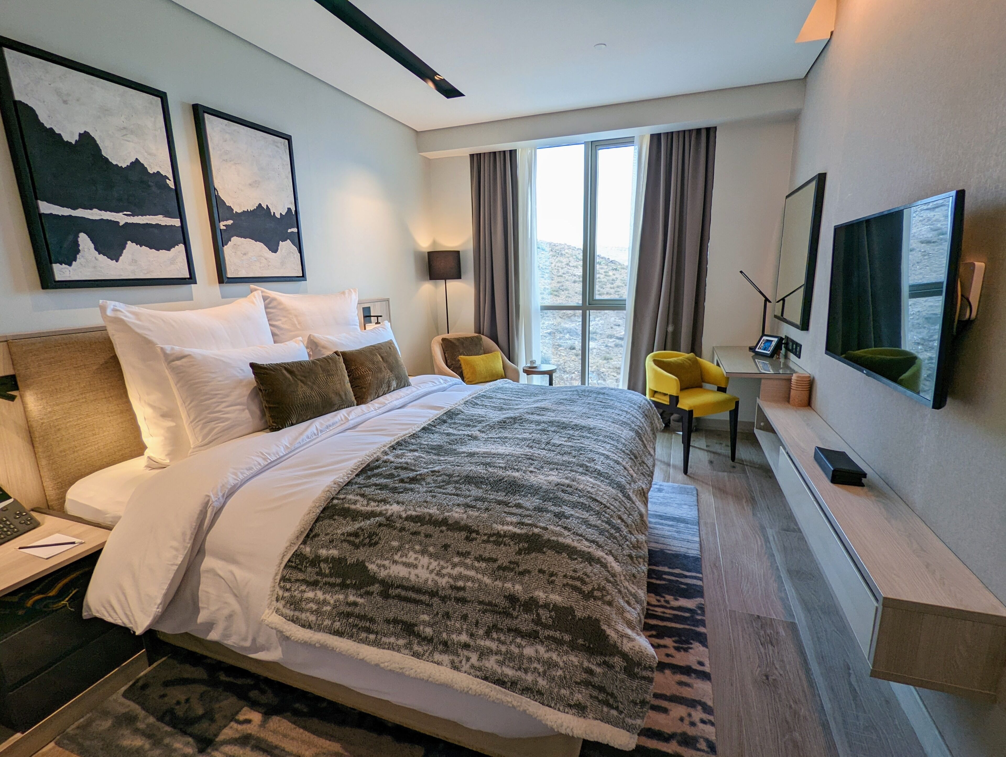 Una habitación de hotel moderna con una cama grande, un escritorio con computadora, una silla y un televisor montado en la pared.