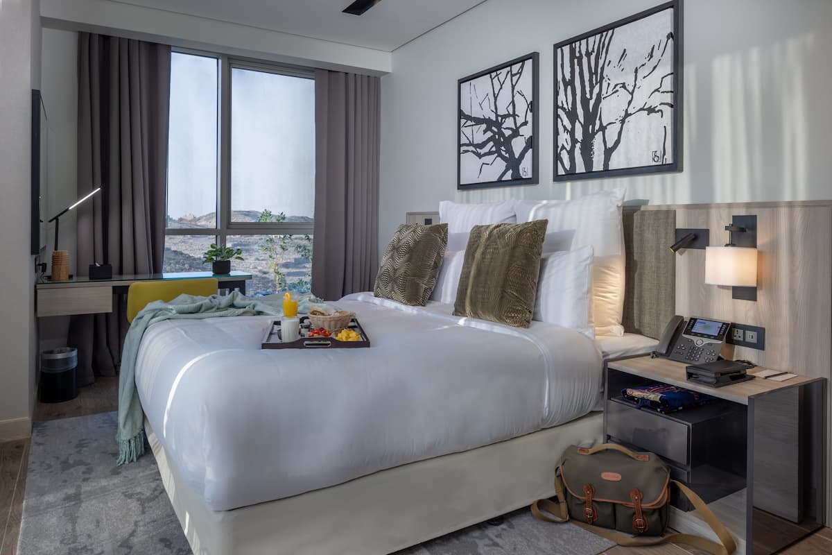 A hotel room with a large bed, a desk with a computer, a bedside table with a lamp, and artwork on the walls.
