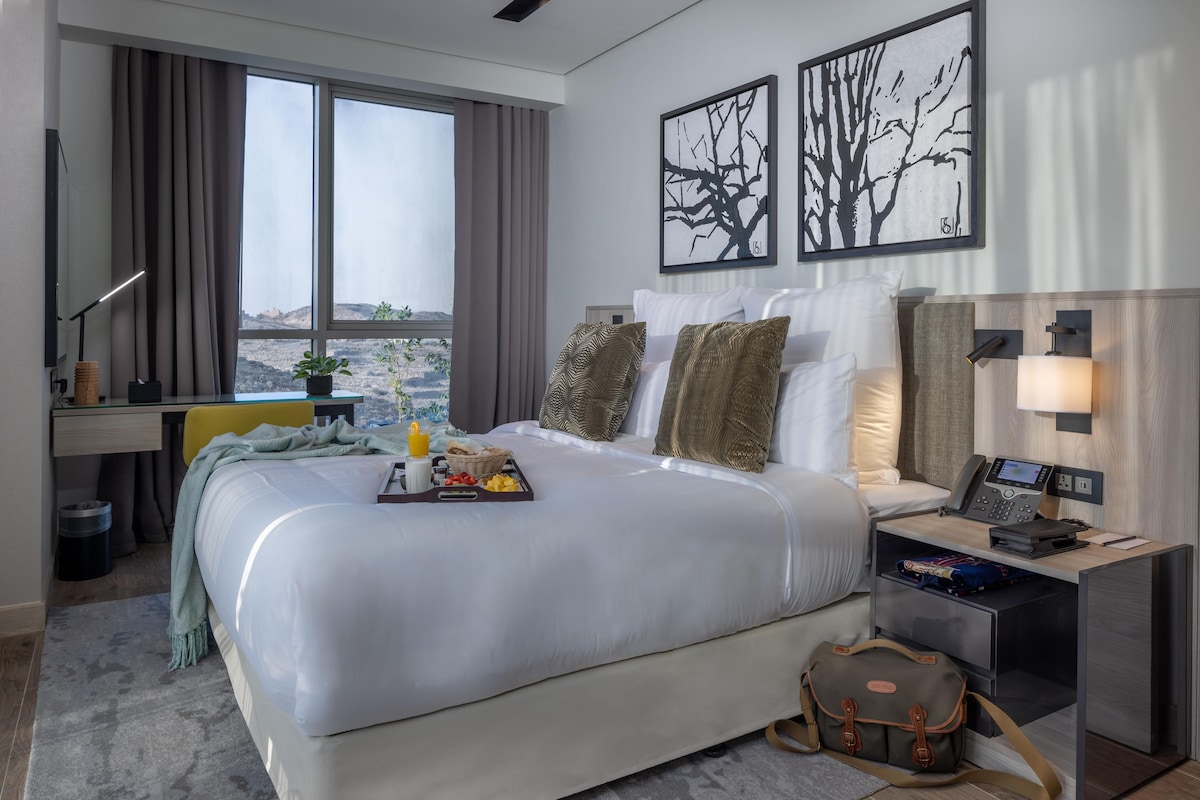 A modern hotel room with a large bed, a desk, a chair, a nightstand with a lamp, and artwork on the walls.
