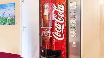 A Coca-Cola vending machine in a hallway with a door and a painting on the wall.