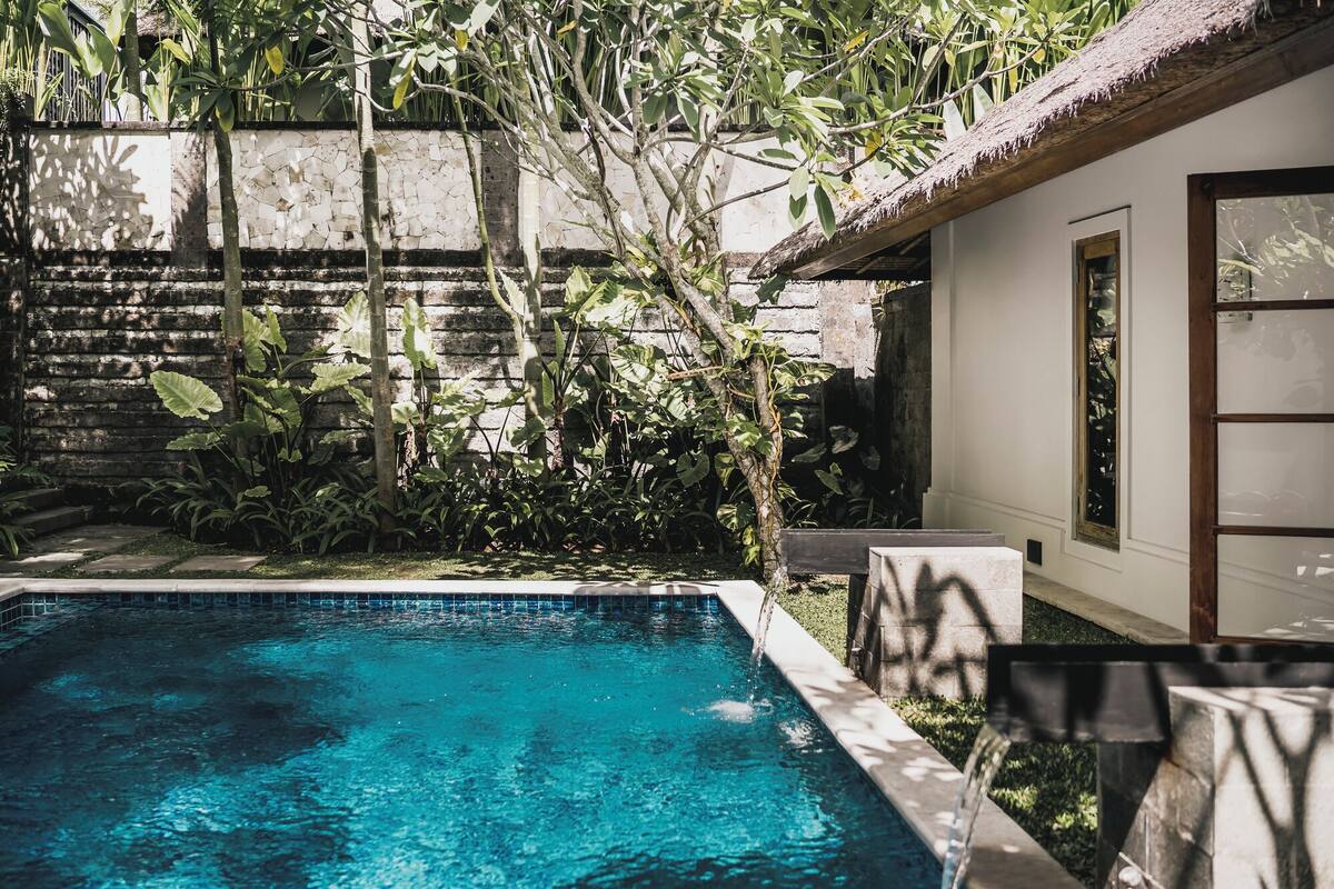 A swimming pool surrounded by lush greenery and a rustic building with a thatched roof.