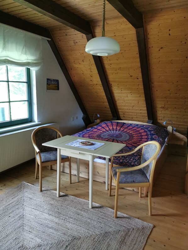 A cozy attic room with a wooden ceiling, a table with chairs, a patterned bedspread, and a hanging light fixture.