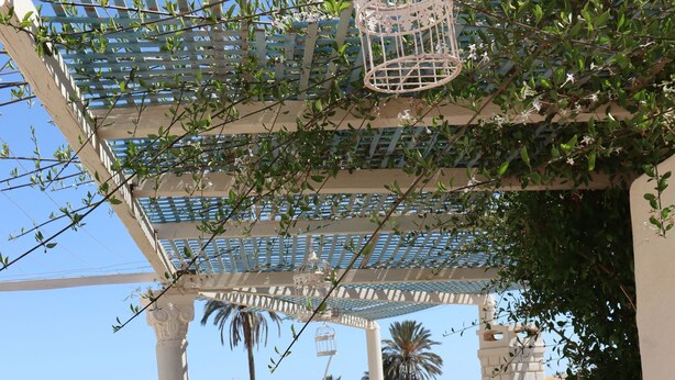 Ein Innenhof mit weißer Pergola, Vogelkäfig und einer Sitzgruppe mit gestreiften Kissen.