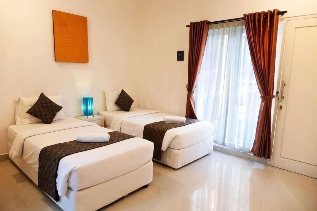 A hotel room with two beds, a nightstand, a lamp, and a sliding glass door with curtains.