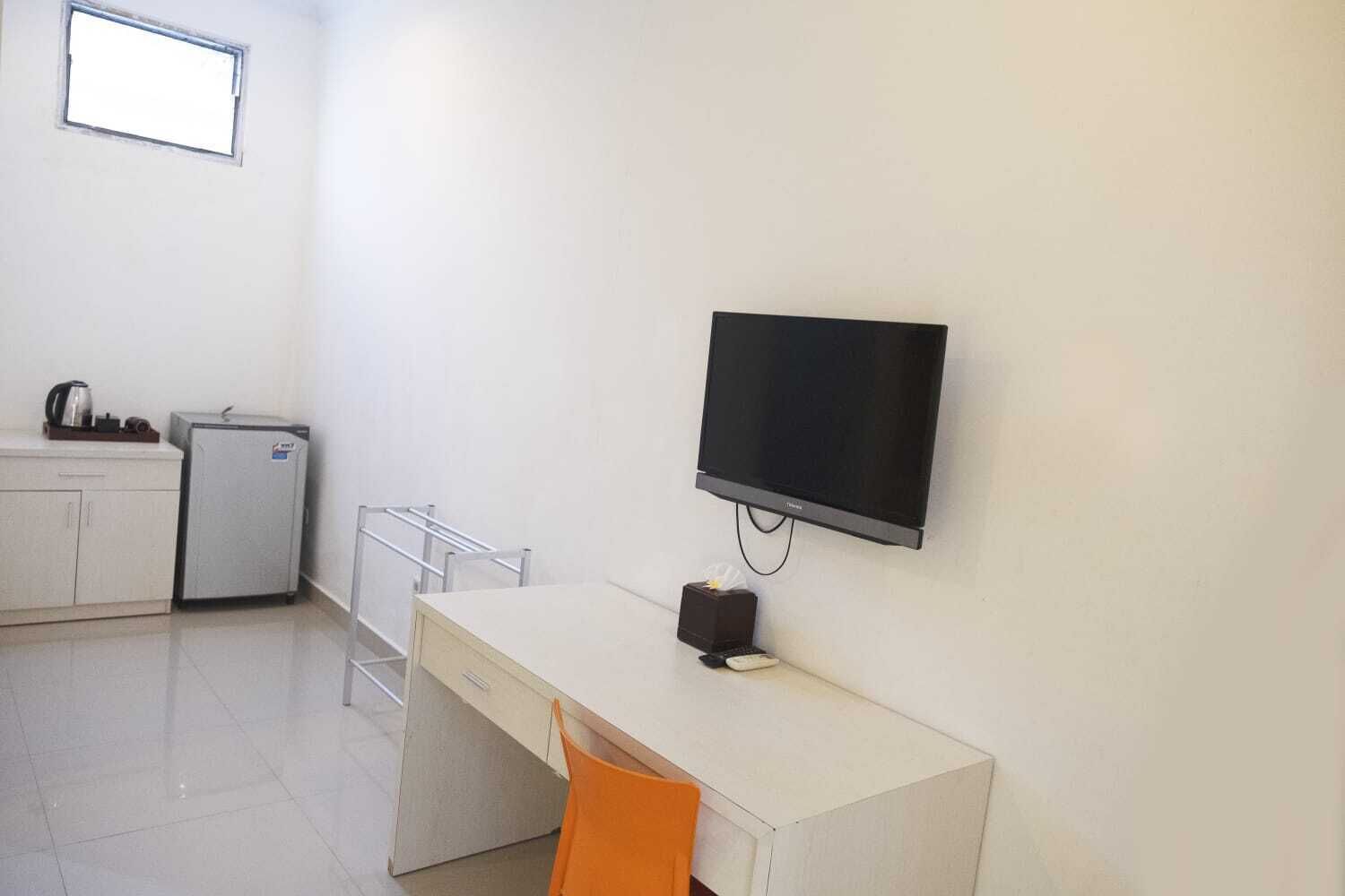 A small, clean room with a desk, chair, television, kitchenette, and a window.