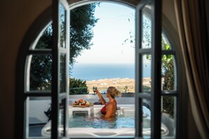 A view from a window of a room with a hot tub, a table with food, and a scenic view of the sea and landscape. - Divino Suites - Adults only (Santorini)