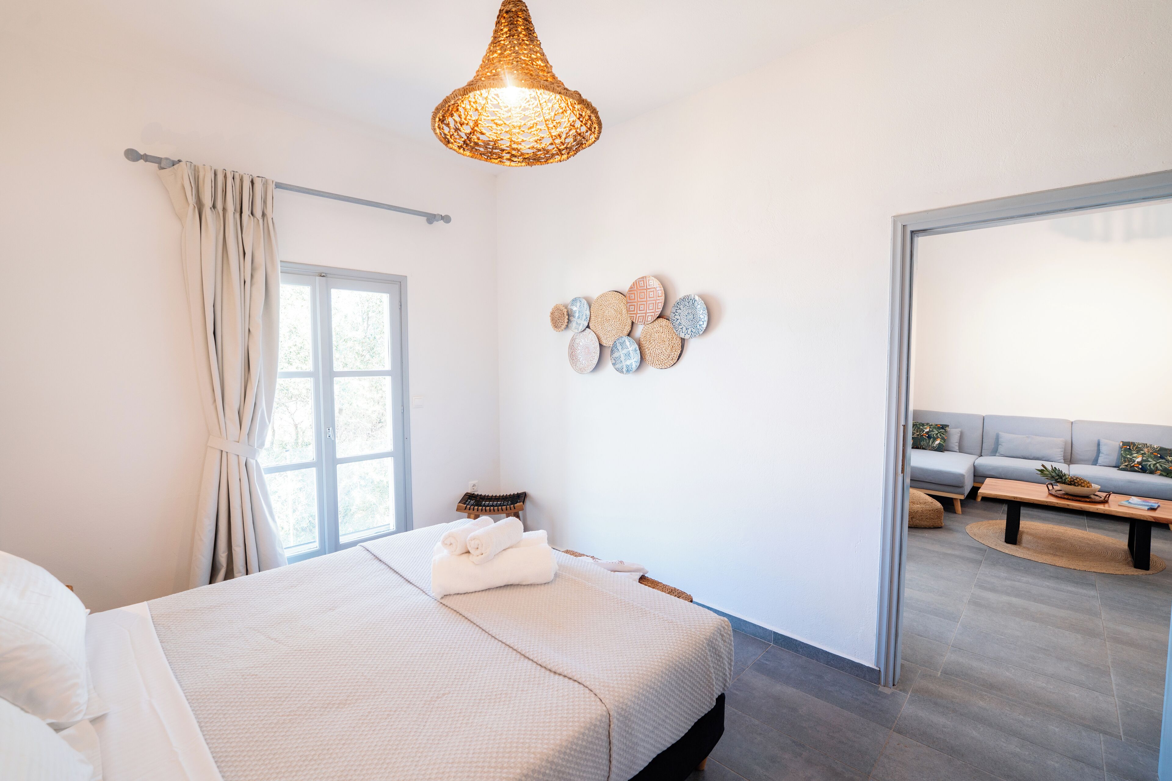 A modern bedroom with a large bed, a hanging light fixture, a window with curtains, and a view of a living area.