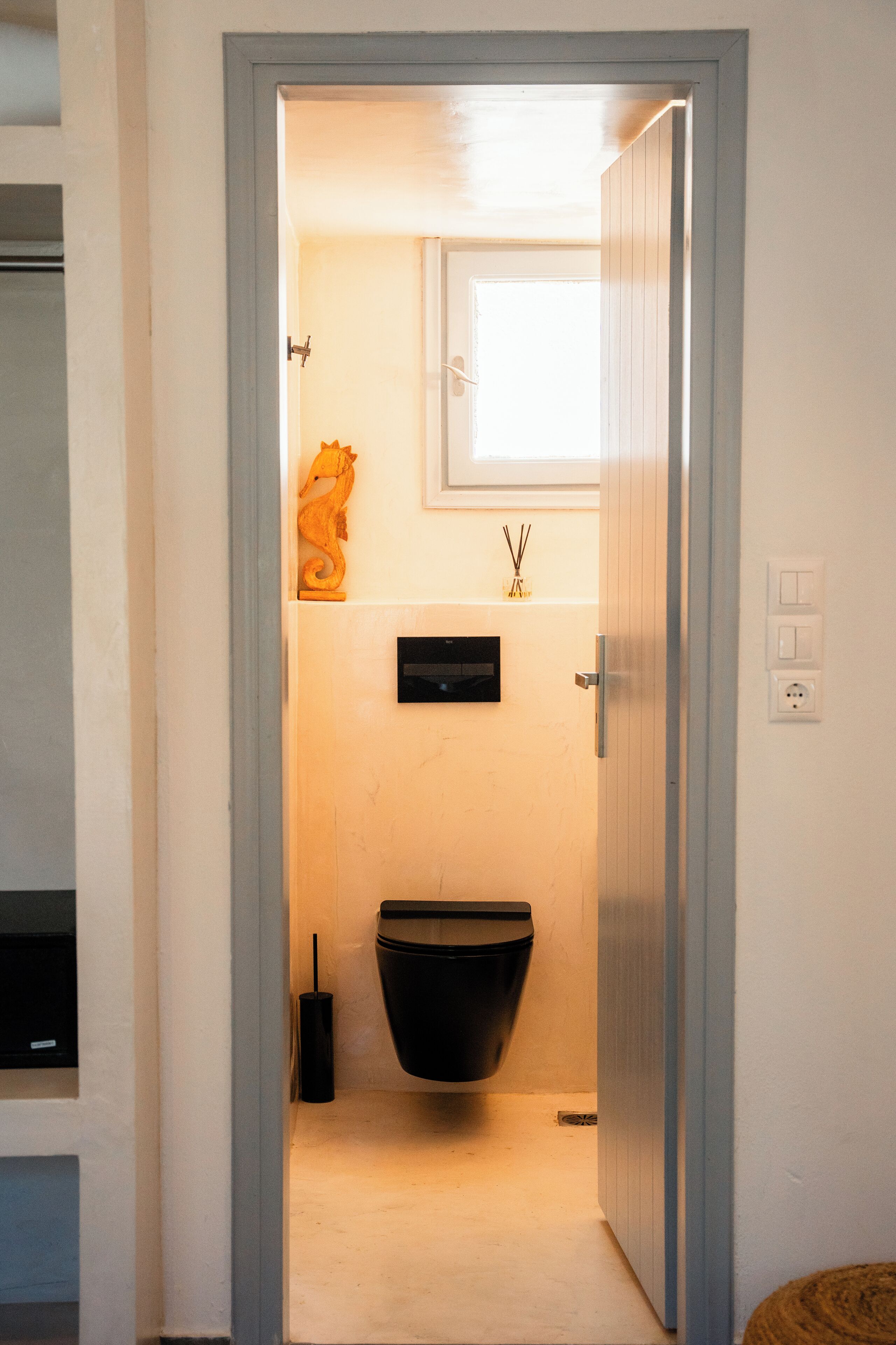Un bagno stretto con il water nero, una piccola finestra e uno scaffale a parete con un oggetto decorativo.