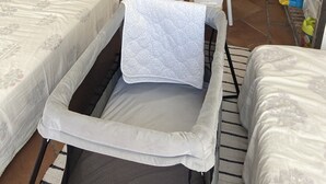 Une petite pièce bien éclairée, avec un berceau, deux lits simples, une petite table et une chaise. On aperçoit une armoire et une porte au fond de la pièce.