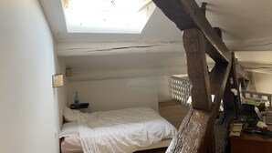 Une chambre avec un puits de lumière, un lit, un bureau avec un ordinateur et une petite table sur laquelle se trouve une bouteille.