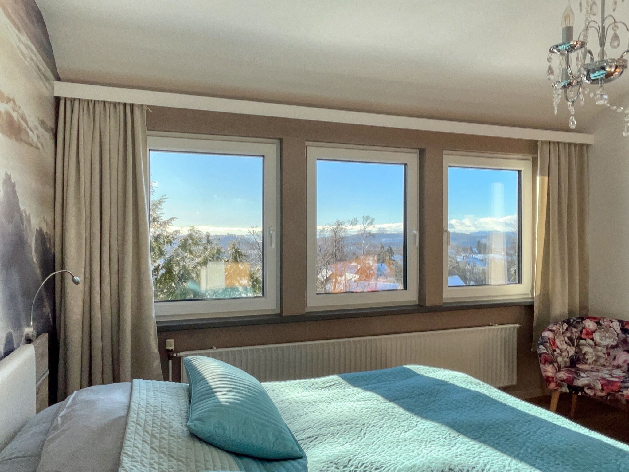 A bedroom with a bed, a chair, and three windows offering a view of a snowy landscape.