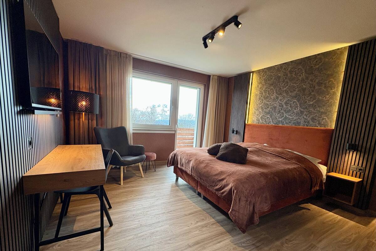 Une chambre d’hôtel moderne avec un plancher en bois, un lit doté d’une tête de lit marron, un bureau avec une chaise et une grande fenêtre avec des rideaux.