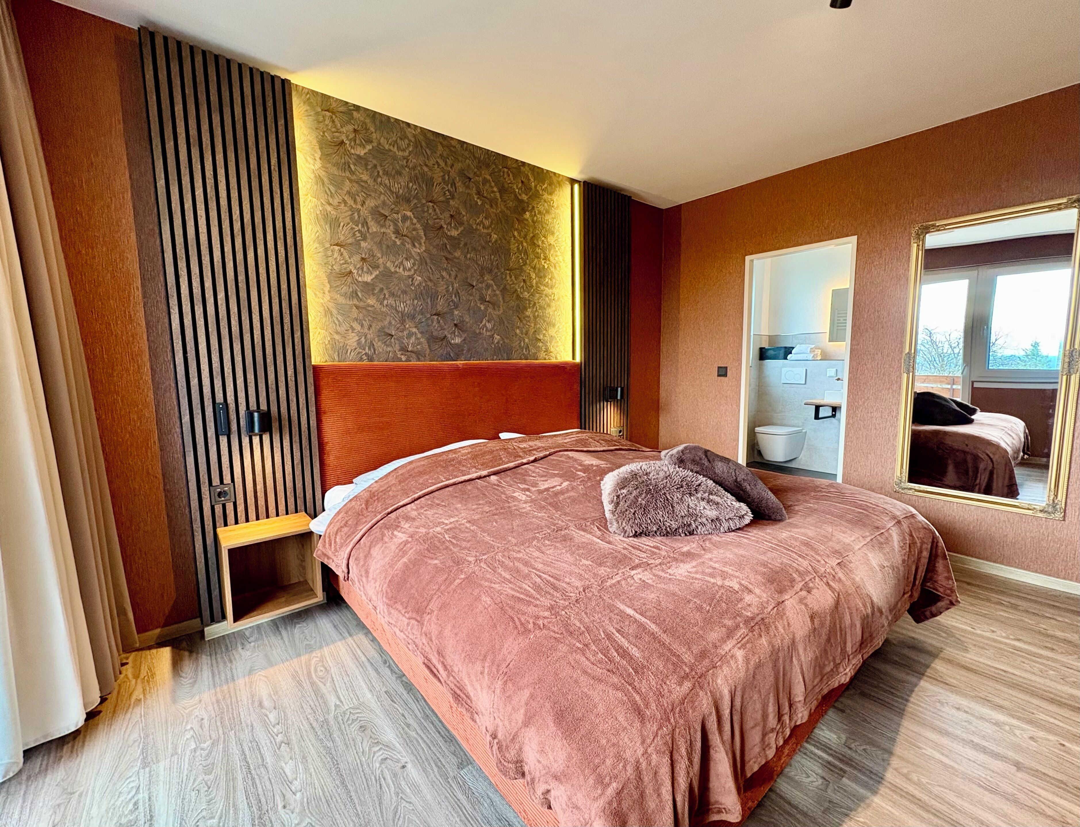 Ein Schlafzimmer mit einem großen Bett, einem Holzkopfende, einem Spiegel und Blick ins Badezimmer.