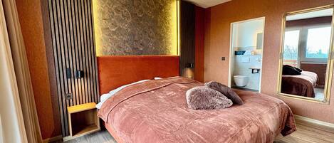 Ein Schlafzimmer mit einem großen Bett, einem Holzkopfende, einem Spiegel und Blick ins Badezimmer.