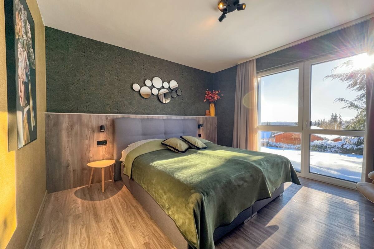 Une chambre moderne avec un grand lit, un plancher en bois, un miroir fixé au mur et une vue sur un paysage enneigé par de grandes fenêtres.