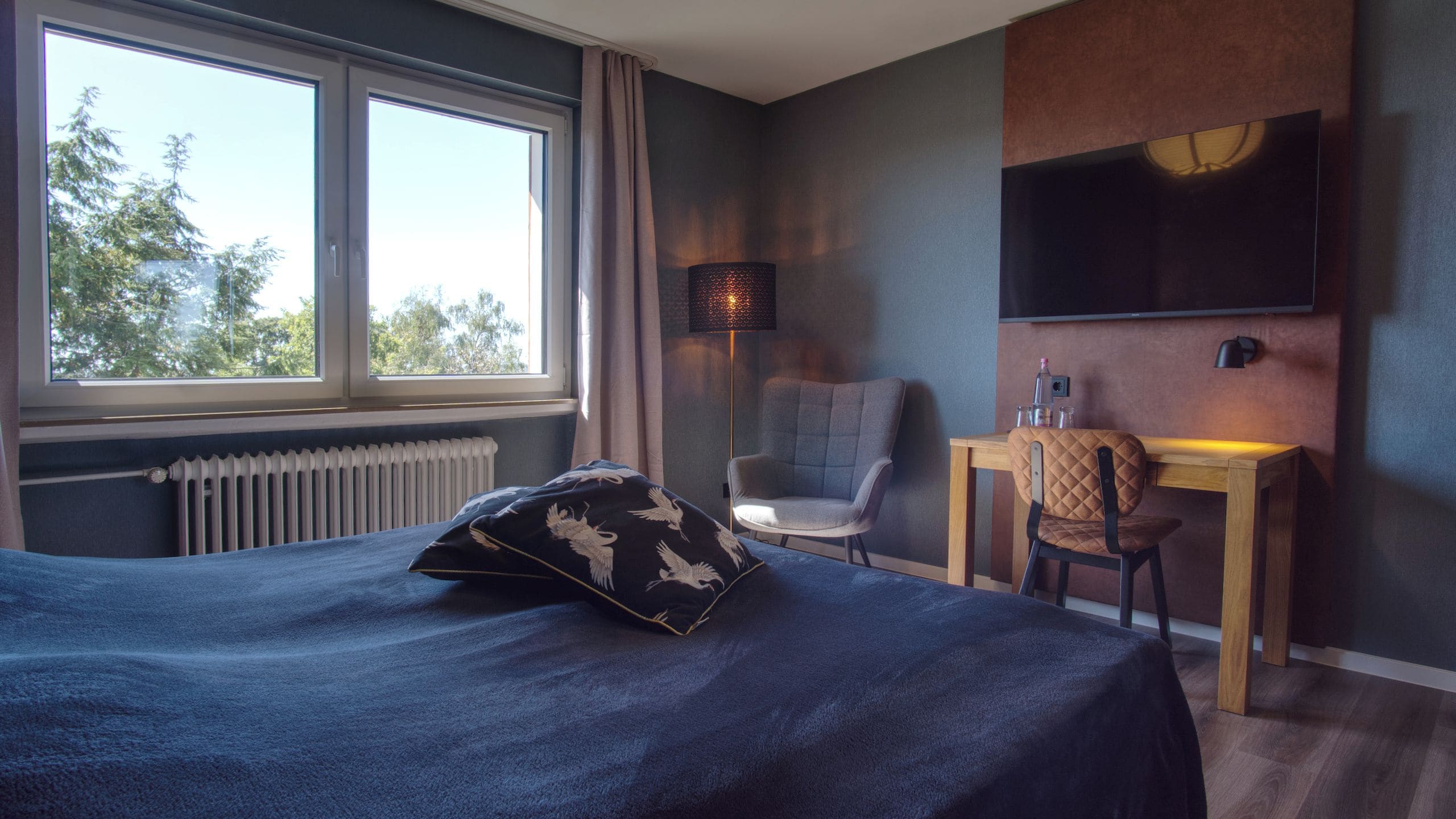 A hotel room with a bed, a desk, a chair, a TV, and a window with a view of trees.