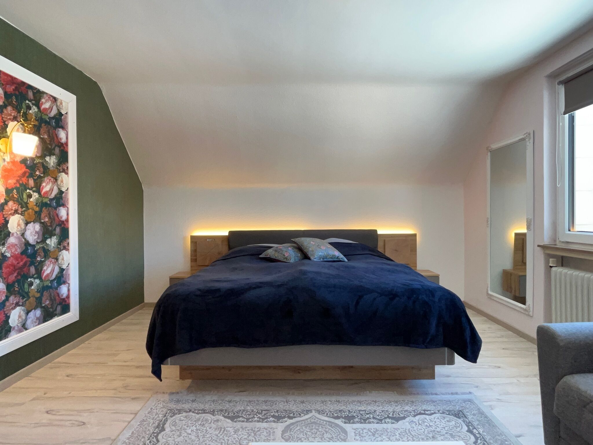 A bedroom with a bed centered under a sloped ceiling, flanked by a large floral painting and a mirror.
