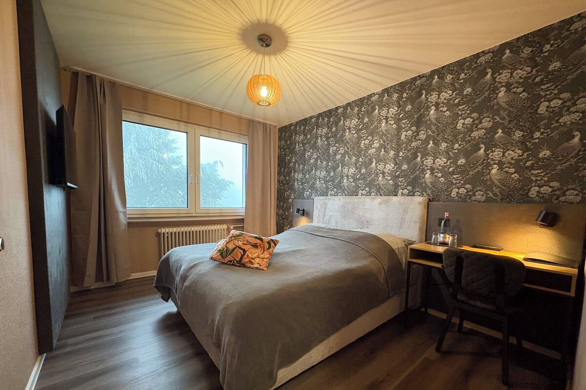 Une chambre à coucher avec un lit, un bureau et une chaise. Il y a une fenêtre avec des rideaux, un radiateur et un mur orné d’un motif floral.