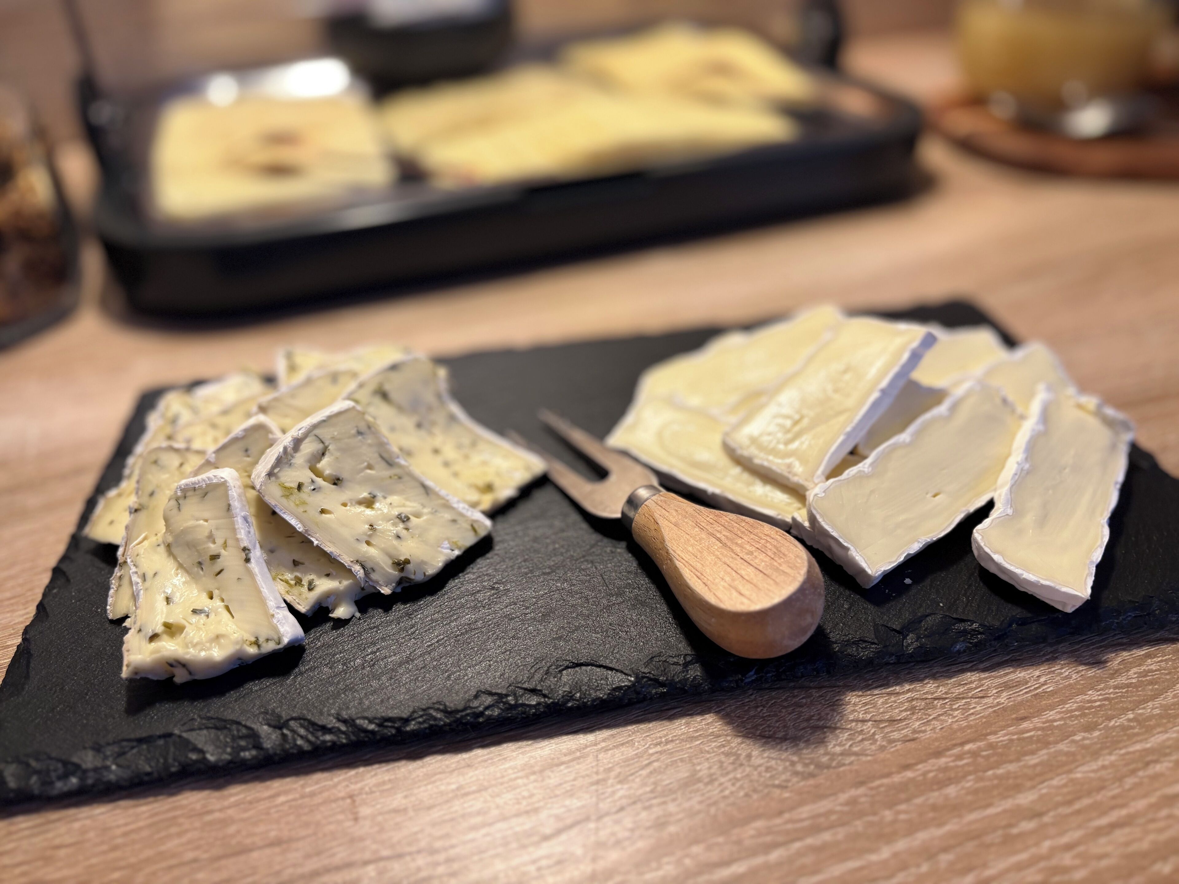 A black slate board with cheese slices and a wooden cheese knife.