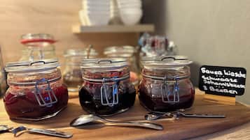Three glass jars with metal clasps on a wooden board, each containing a dark red liquid, accompanied by a spoon. A sign in the background indicates the contents are Sauerkirsche, Schwarzwälder Kirschen, Johannisbeers, and 3 Beeren.
