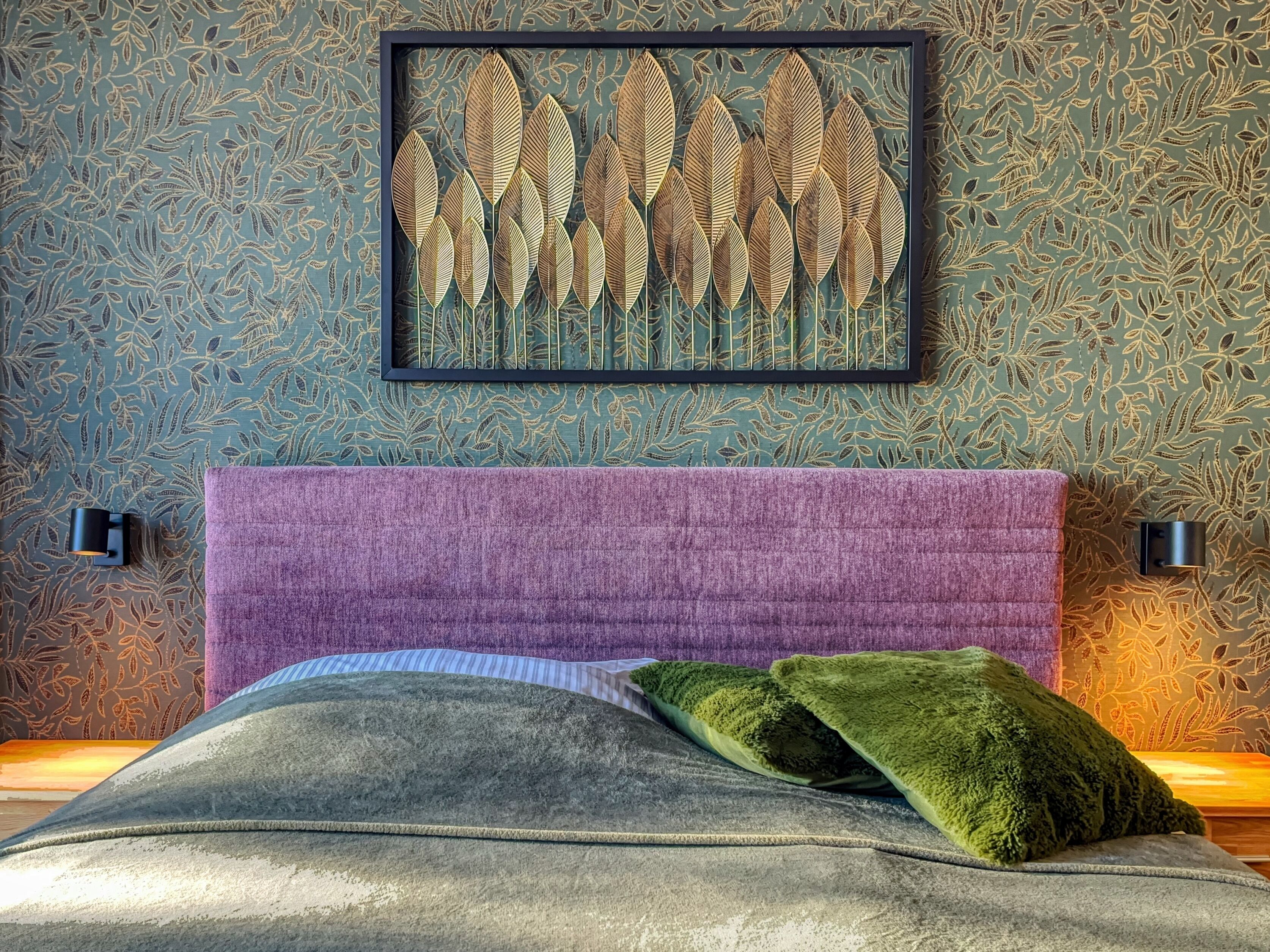 Un dormitorio con cabecera morada, una obra de arte con diseño de hojas sobre la cama y un cojín decorativo verde.