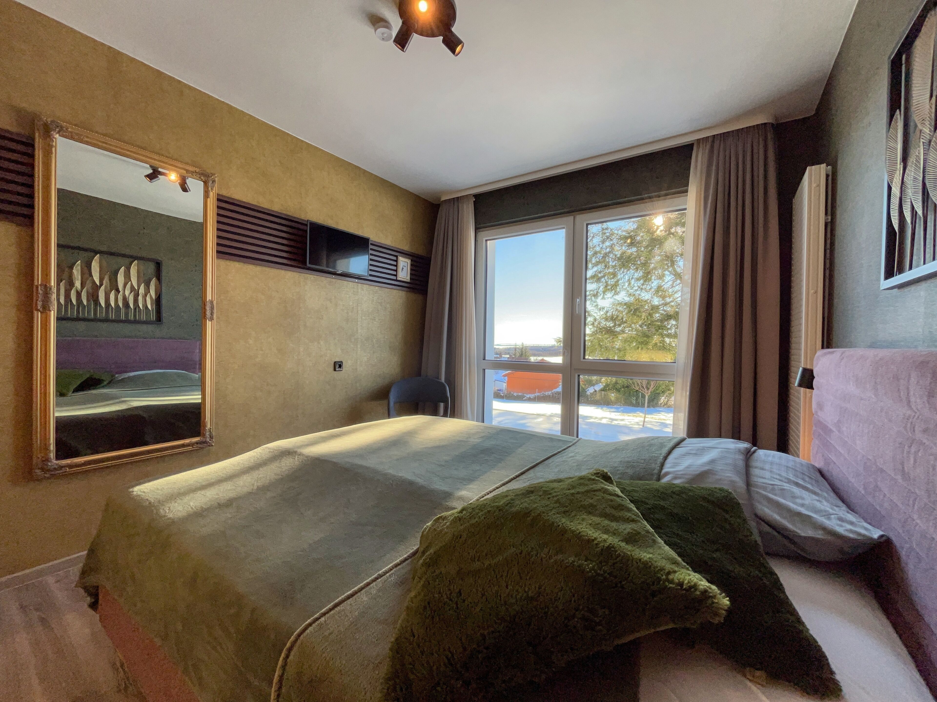 A bedroom with a bed, a large mirror, a window with curtains, and a view of the outside.