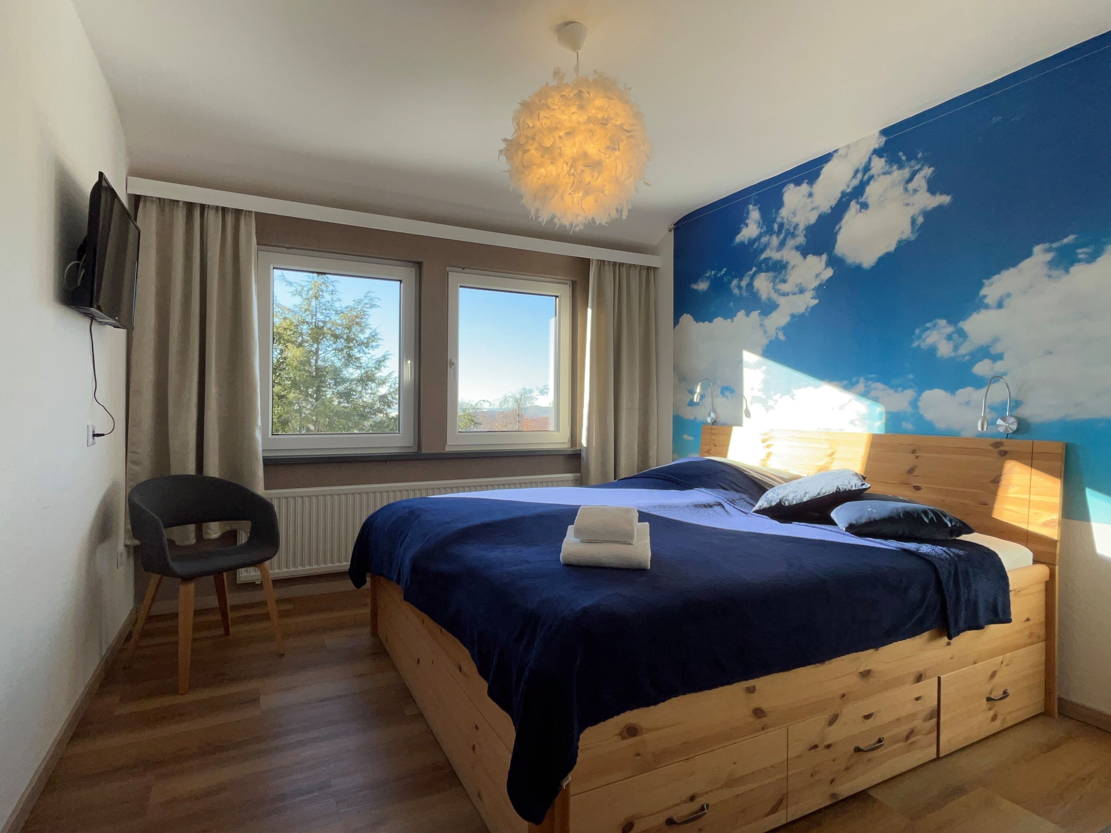Un dormitorio con cama, cabecera de madera, una silla, un televisor y una ventana con cortinas.